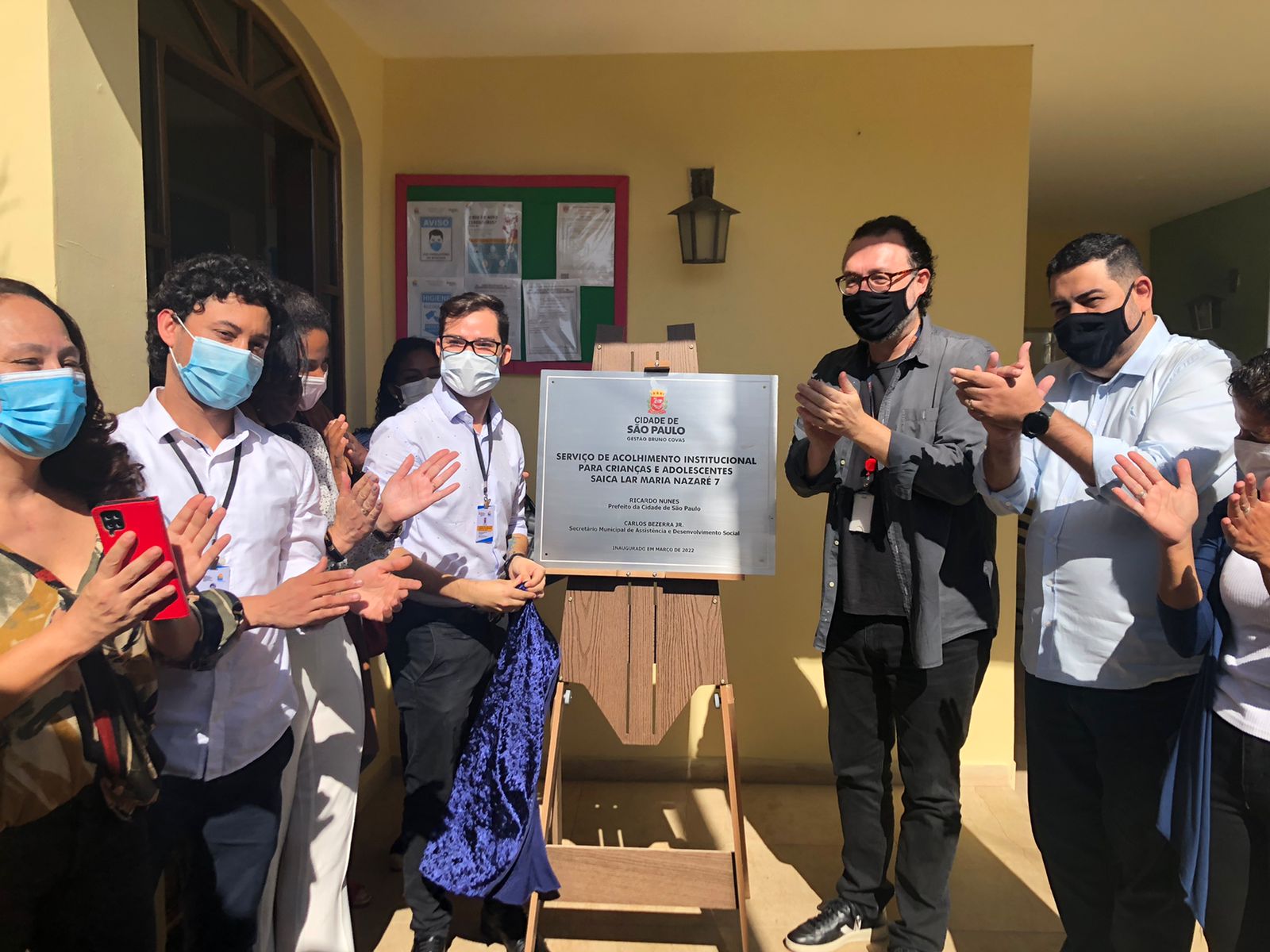 Cinco pessoas do lado esquerdo lado a lado batendo palmas, no meio um cavalete com placa de metal escrito Serviço de Acolhimento Institucional para Crianças e Adolescentes Saica Lar Maria de Nazaré 7 e do lado esquerdo secretário ao lado do subprefeito.
