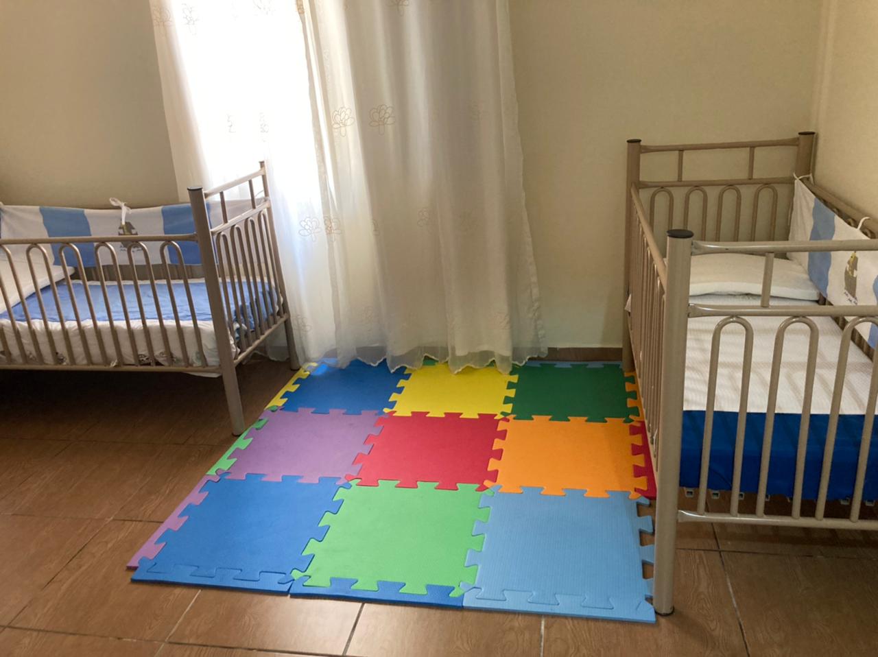 Dois berços de estrutura metálica, com colchoes cobertos por um lençol azul. No meio um tapete de encaixar colorido.