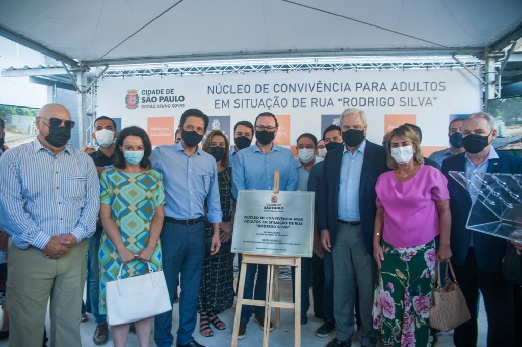 Autoridades estão em pé, sendo cinco homens e duas mulheres, todos de máscara, em primeiro plano, atrás de uma placa de inauguração. Em segundo plano, pessoas em pé acompanham o evento.