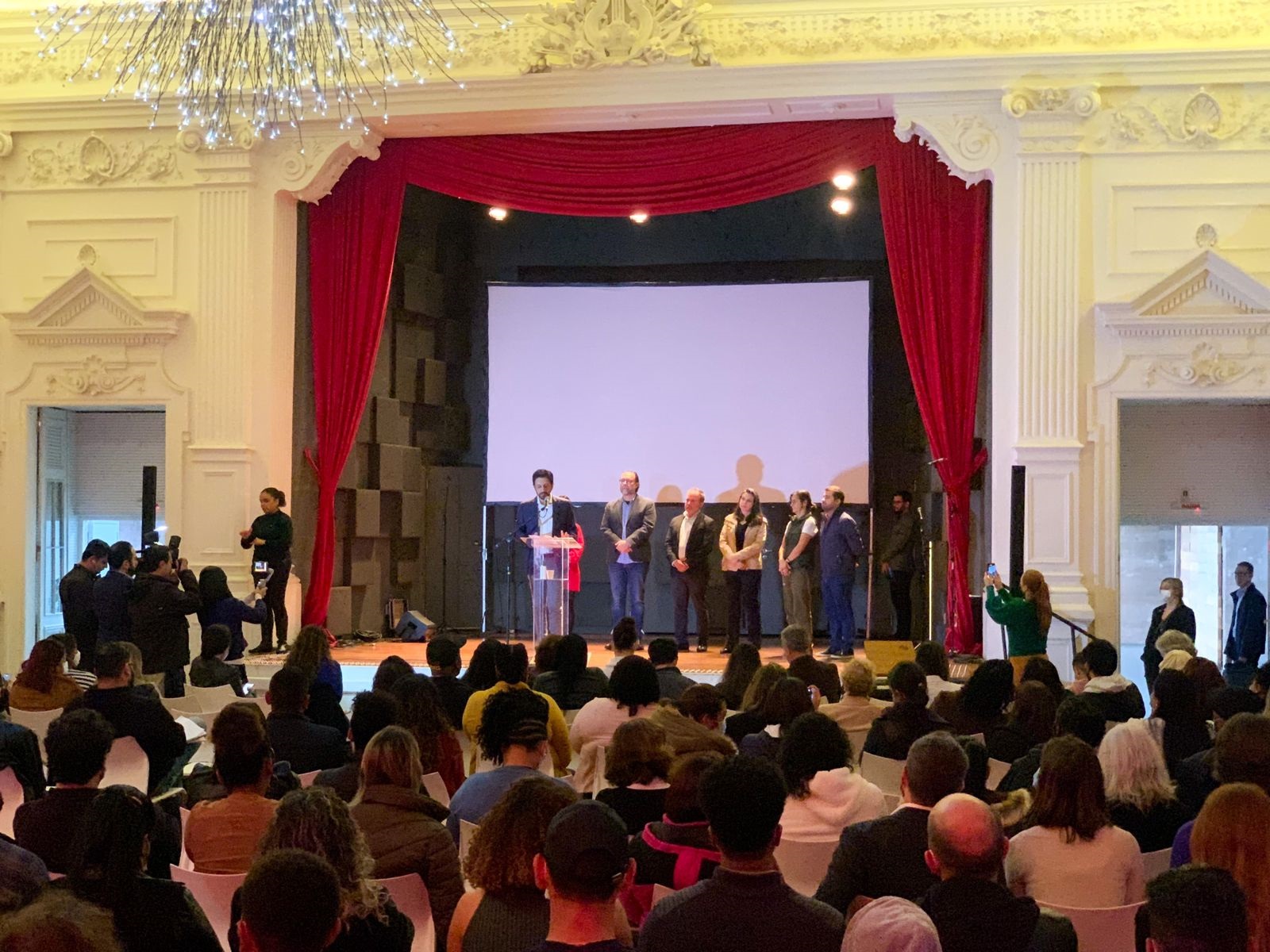 Prefeito Ricardo Nunes em um púlpito de acrílico em cima do palco falando ao microfone. Ao lado dele o Secretário de Assistência Social Carlos bezerra Jr. e outras pessoas ao fundo. Dezenas de pessoas estão na plateia, todas de costas.