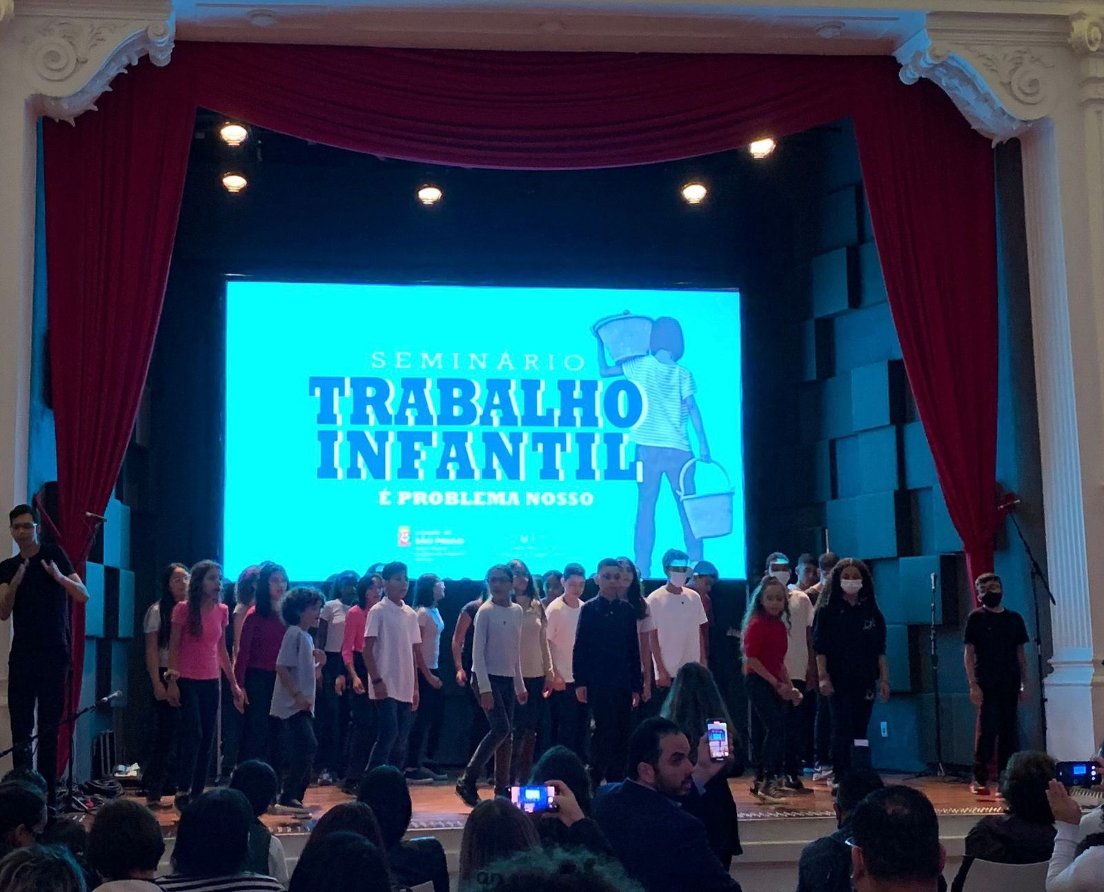 Telão com fundo azul e os dizeres: Trabalho Infantil é problema nosso!. A frente, crianças do Coral Infantil Bacarelli se apresentam.