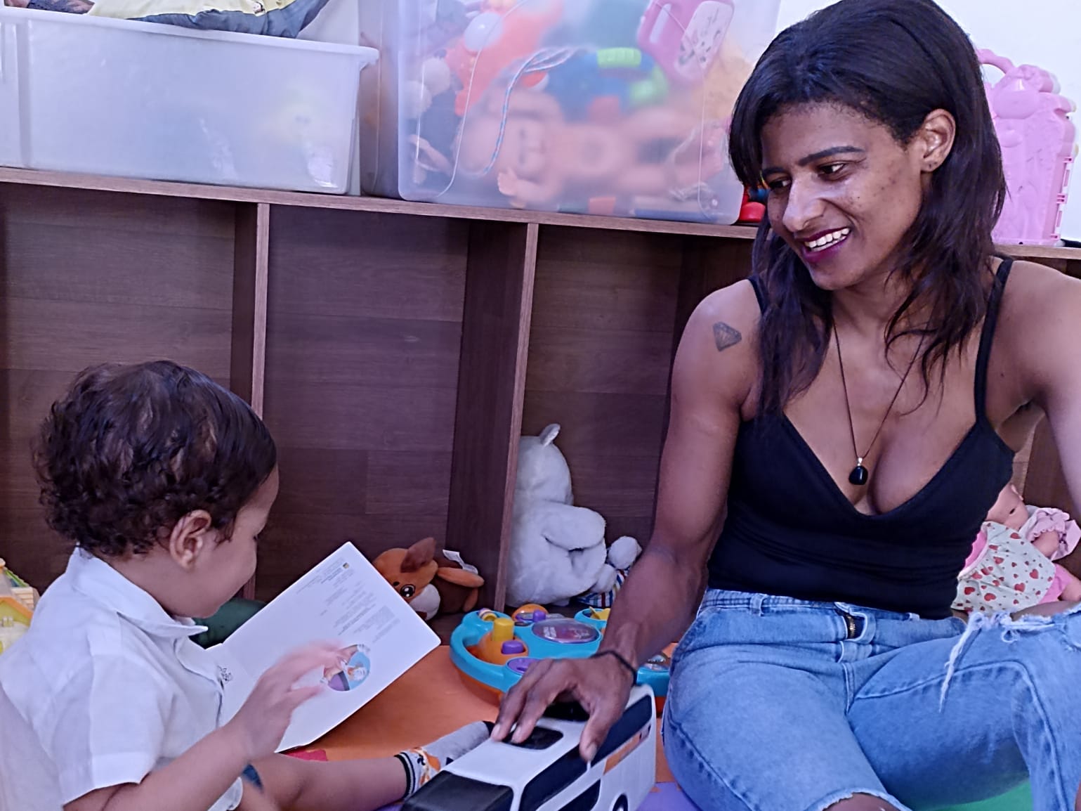 Uma mulher está sentada em um tapete colorido com seu filho pequeno. Ela veste uma regata preta e uma calça Jeans, enquanto a criança usa uma camiseta branca e uma bermuda. O bebê brinca com um ônibus de brinquedo em um livro azul com outros brinquedos por perto.