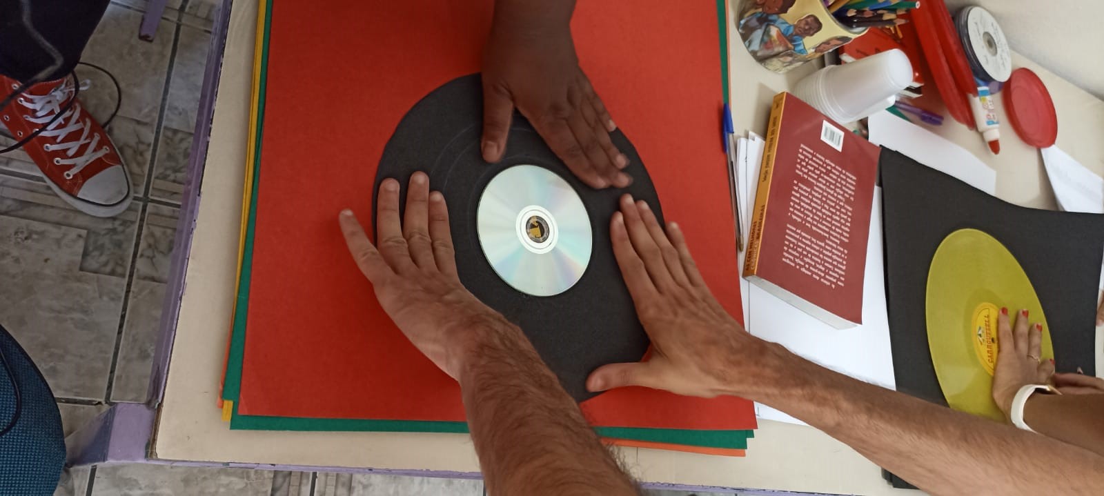 Em uma mesa coberta por um tecido vermelho, duas mãos seguram um disco de vinil confeccionado com papel preto e um cd no meio. A mão de outra pessoa também segura o disco.