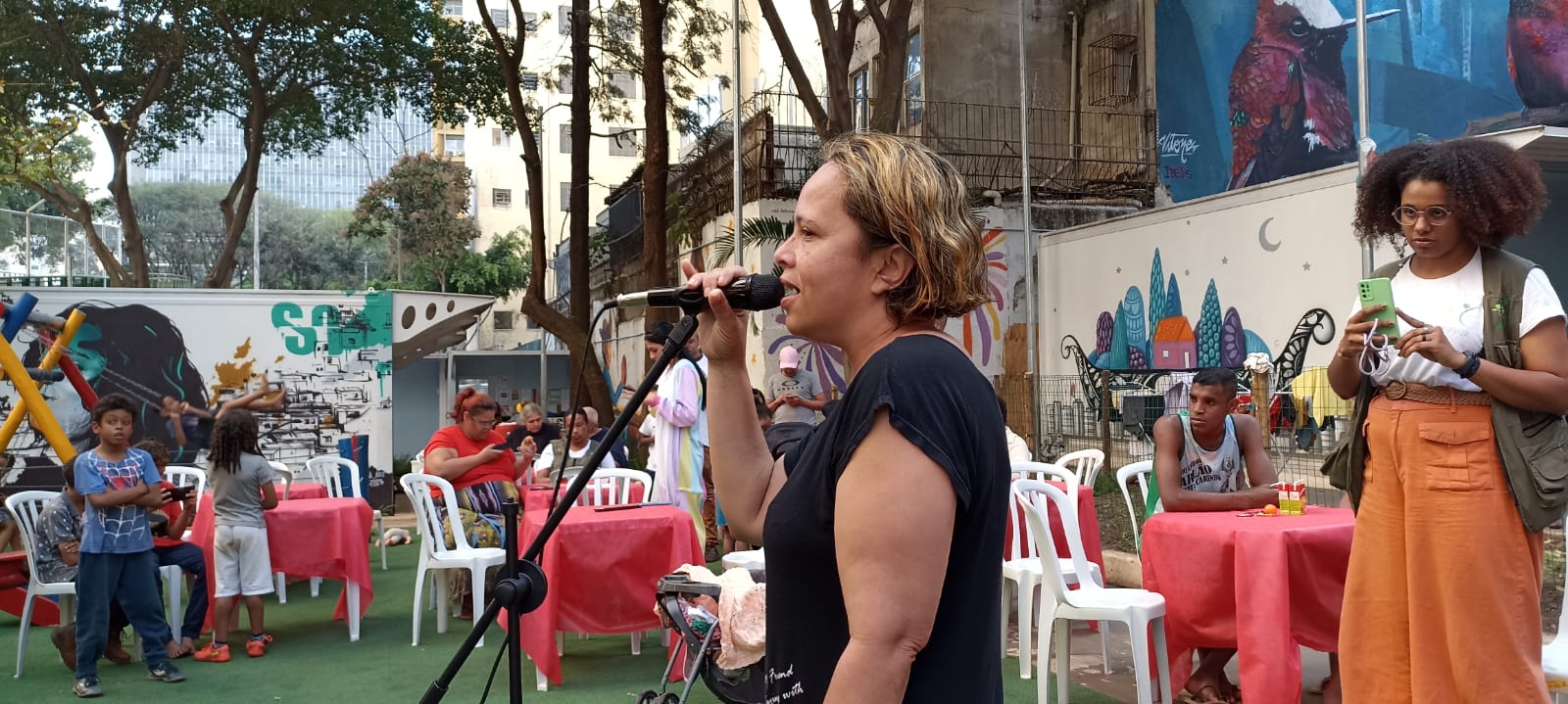 Uma mulher, vestindo uma blusa e uma camiseta preta, está na frente de um microfone com um tripé, cantando. Elas têm cabelos curtos até os ombros com mechas loiras.
