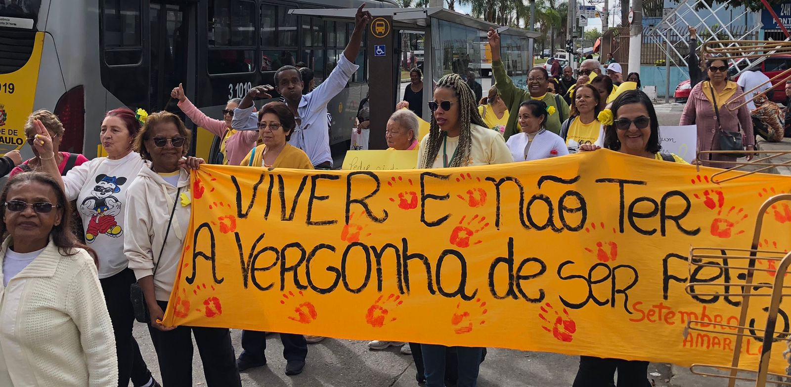 Varias pessoas vestindo roupas amarelas caminham pela rua. Elas carregam placas e cartazes pela campanha de setembro amarelo.