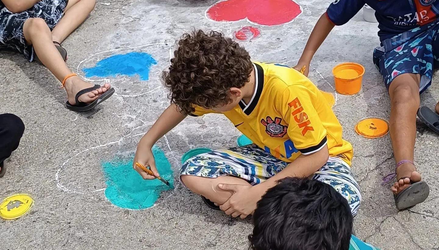 Na imagem, um garoto de cabelos cacheados está pintando um coração verde na calçada. Ele veste uma camiseta amarela do Corinthians e uma bermuda branca estampada. Outras crianças ao redor dele realizam a mesma ação. 