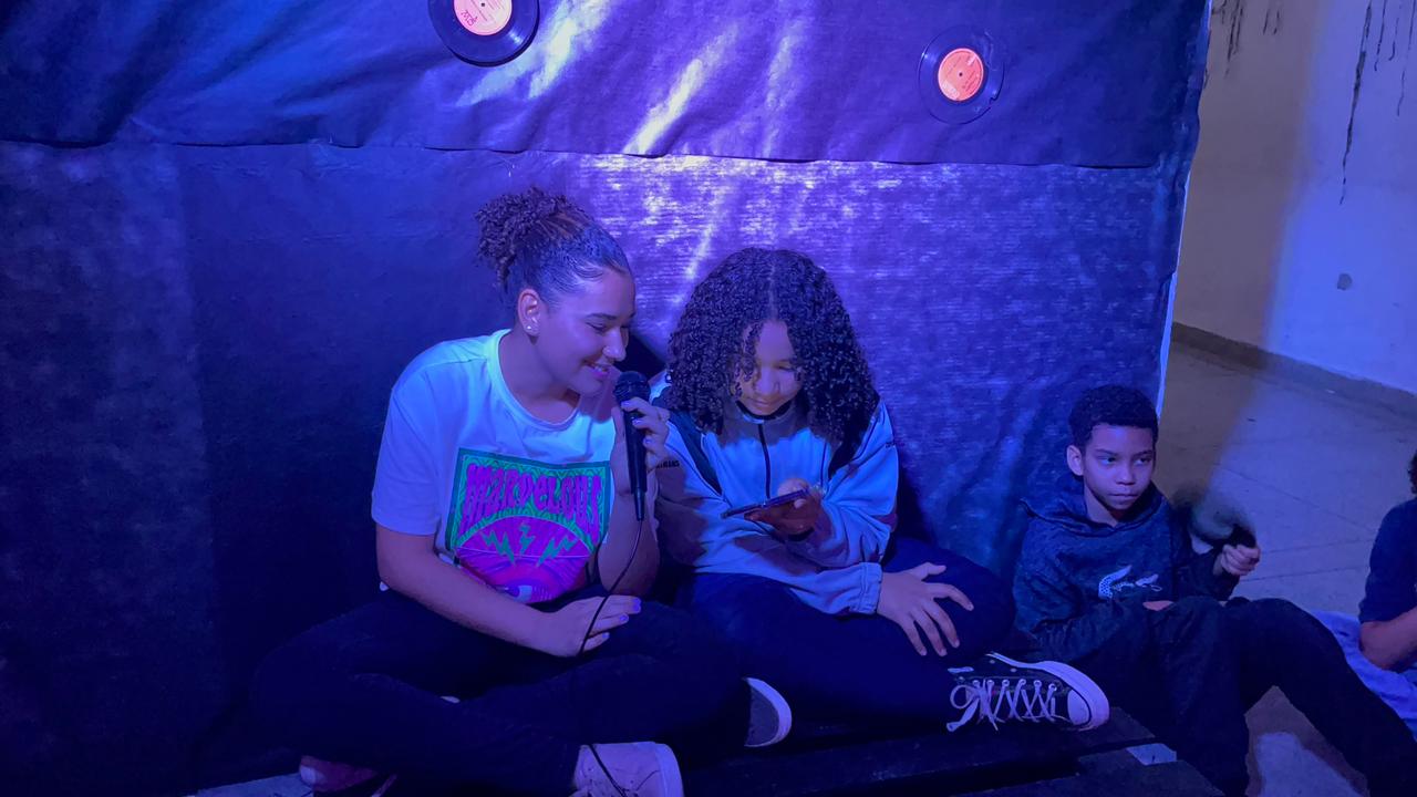 Na imagem, duas meninas estão sentadas no chão uma ao lado da outra. A da direita está com um celular na mão, enquanto a outra segura um microfone. Ambas são iluminadas por uma luz azul.