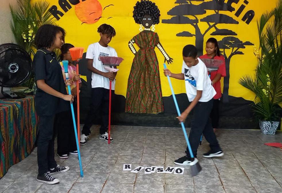 Na imagem, cinco crianças fazem uma apresentação. Todas seguram vassouras nas mãos. Um garoto usa a vassoura para varrer folhas que estão no chão com letras que compõem a palavra “racismo”