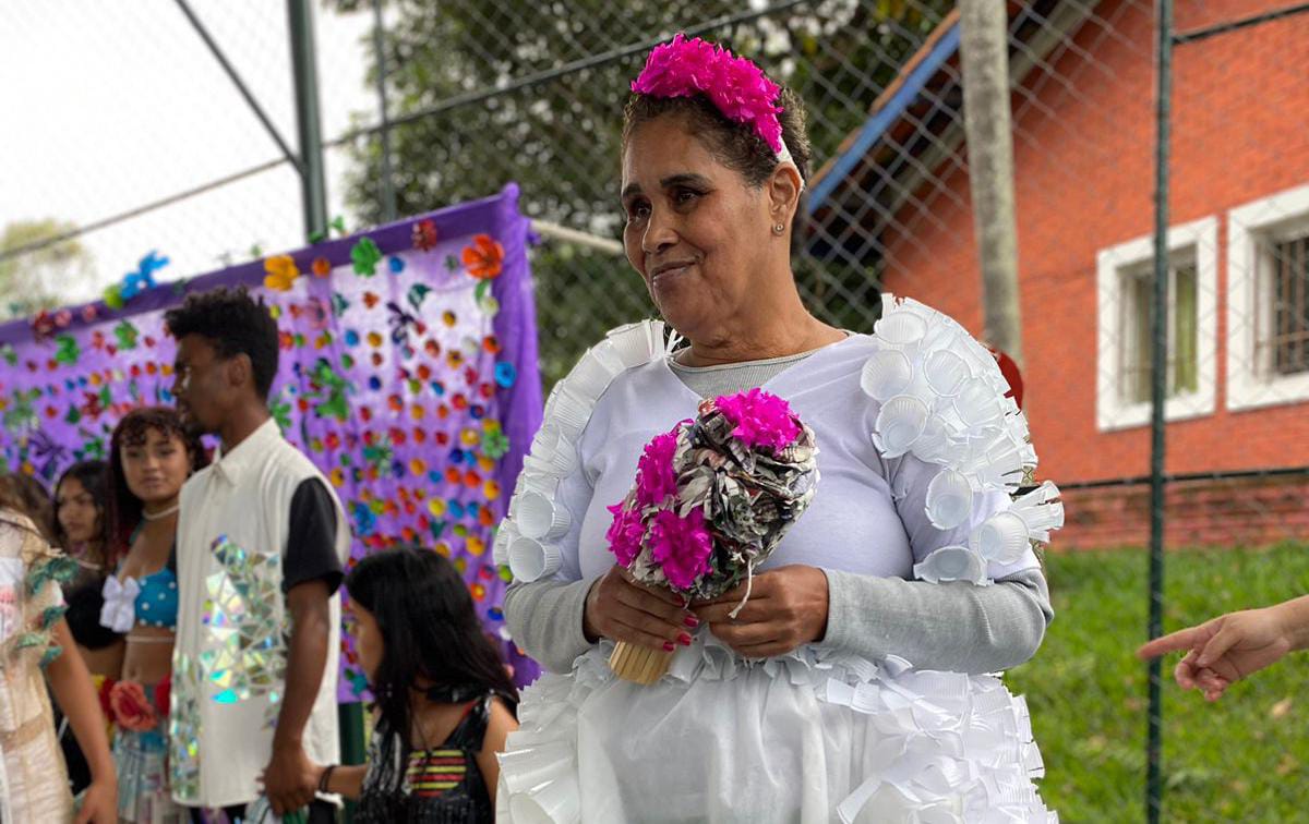 Uma mulher está usando um vestido de noiva branco feito com vários copos descartáveis. Ela segura um buquê feito de papel rosa e folhas de jornais e usa uma tiara de flores de papel rosa. 