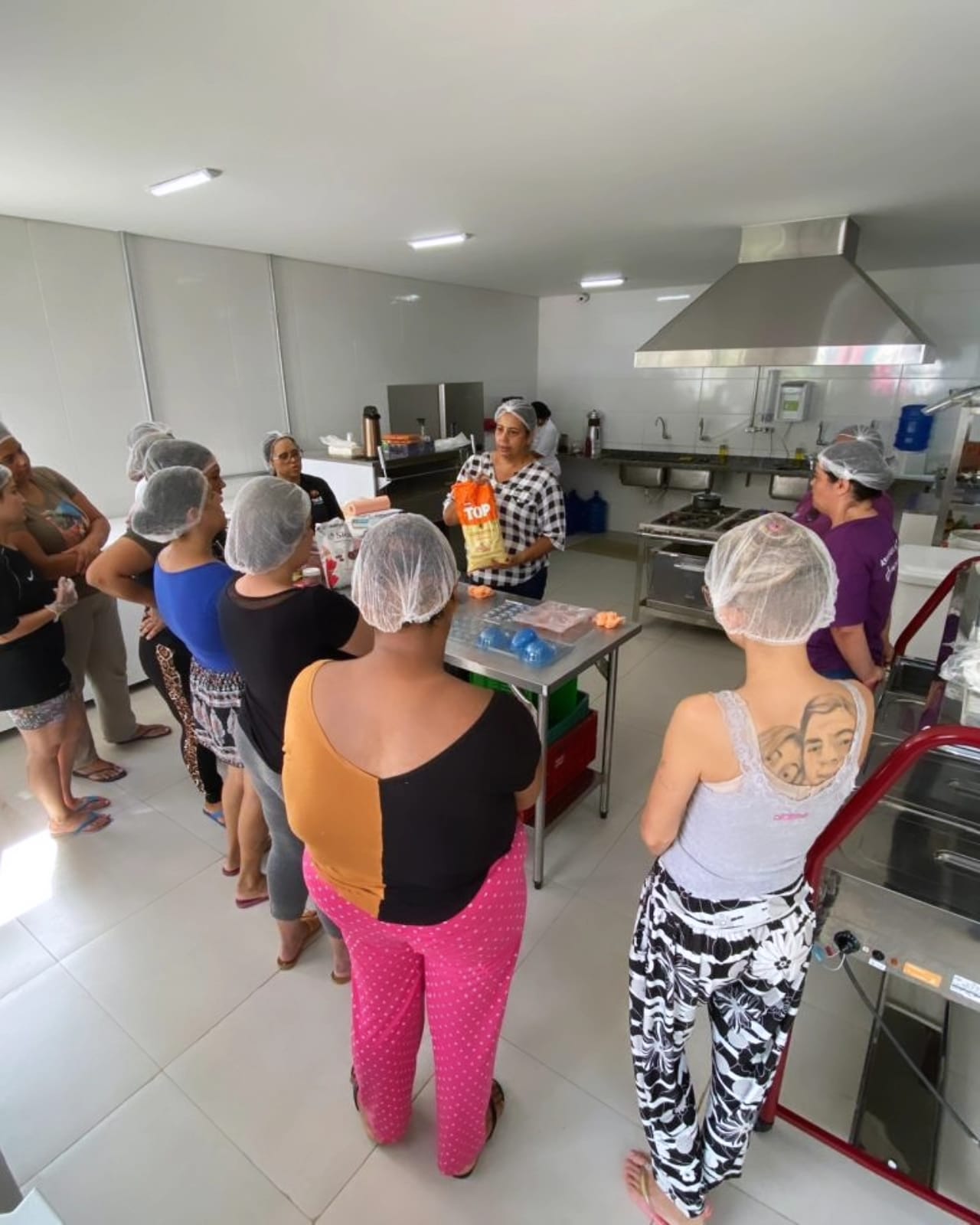Na imagem, moradores da Vila Reencontro Santo Amaro estão reunidos em um círculo na cozinha, trabalhando na confecção de ovos de chocolate. Eles usam luvas e toucas de proteção. Uma mulher está na frente deles, repassando orientações.