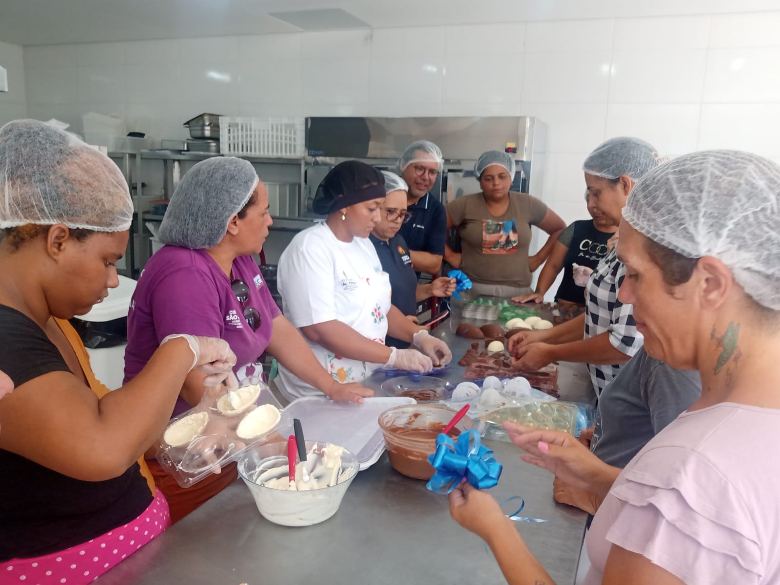 Na imagem, os moradores da Vila Reencontro estão reunidos na cozinha, trabalhando na confecção de ovos de chocolate. Eles usam luvas e toucas de proteção. Há vários utensilios na mesa, como formas e vasilhas com chocolate derretido.