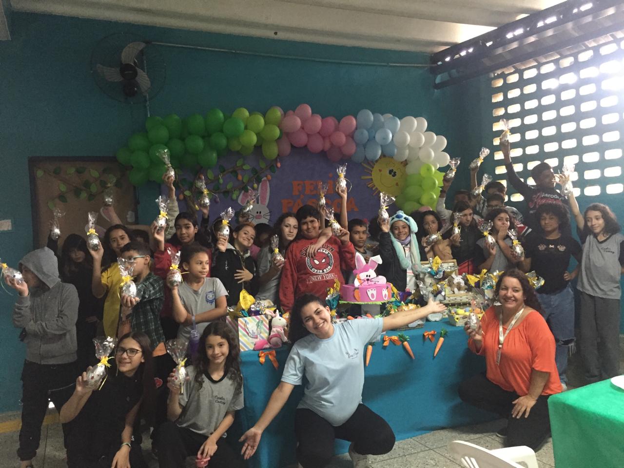 Na imagem, a equipe do CCA, a ex-atendida Anna Julia e crianças se juntam para uma foto no local decorado.Todos os atendidos seguram ovos de Páscoa. 