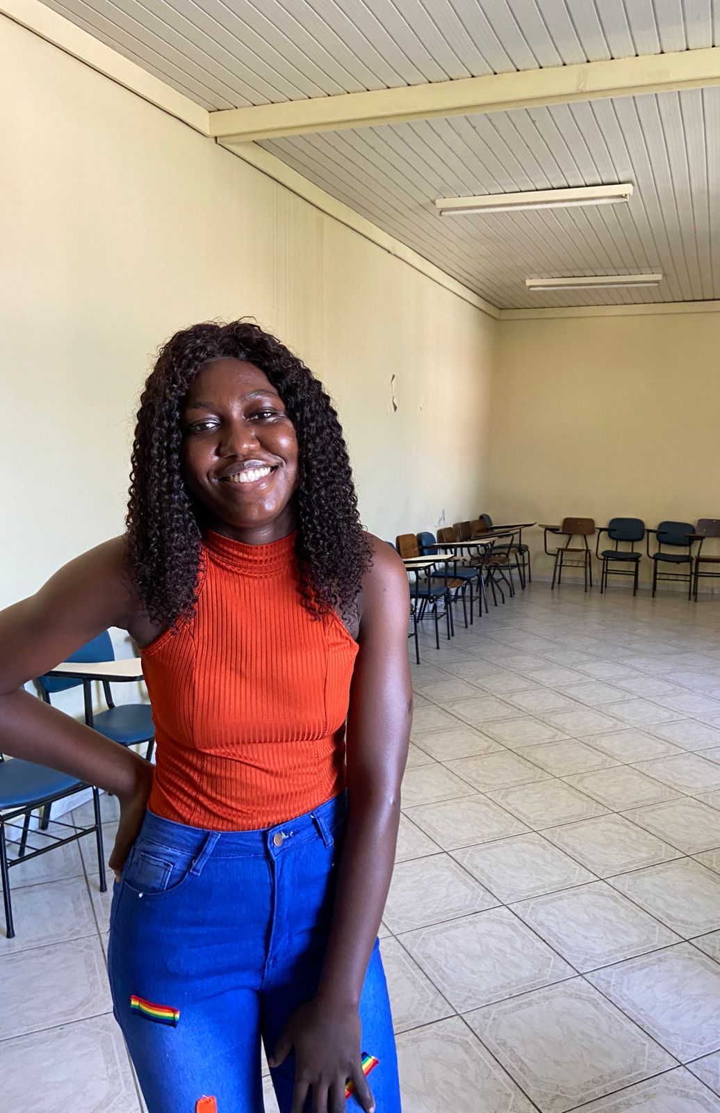 Em uma sala com carteiras de estudo ao redor, uma mulher vestindo camiseta laranja e calça jeans posa para foto.