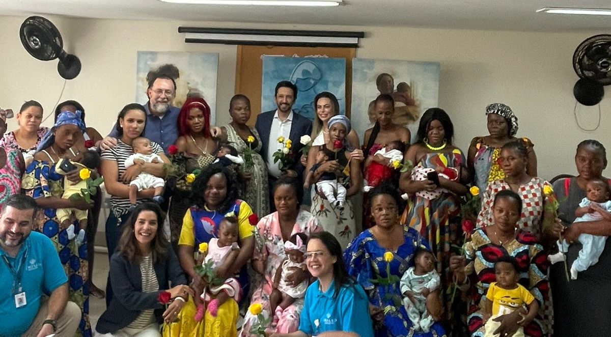 Em frente de uma parede branca, com dois ventiladores pretos e quadros, pessoas posam para foto sorrindo. Há 16 mulheres, das quais 13 seguram bebês em seus colos. Todas vestem roupas coloridas, e algumas tem véus em seus cabelos. Há 2 funcionários, uma mulher e um homem brancos, vestindo camisetas azuis, com o nome da Organização da Sociedade Civil. 