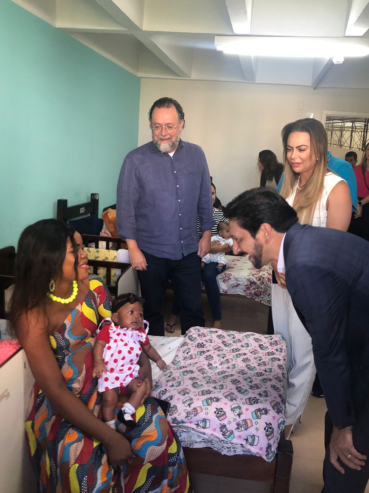 Uma mulher negra, com vestido colorido, está sentada numa cama de madeira escura, e segura em seu colo uma bebe negra, com vestido branco com bolhinhas vermelhas. Ela olha para um homem branco, vestido com camisa branca e terno azul marinho, que se abaixa para falar com a mulher