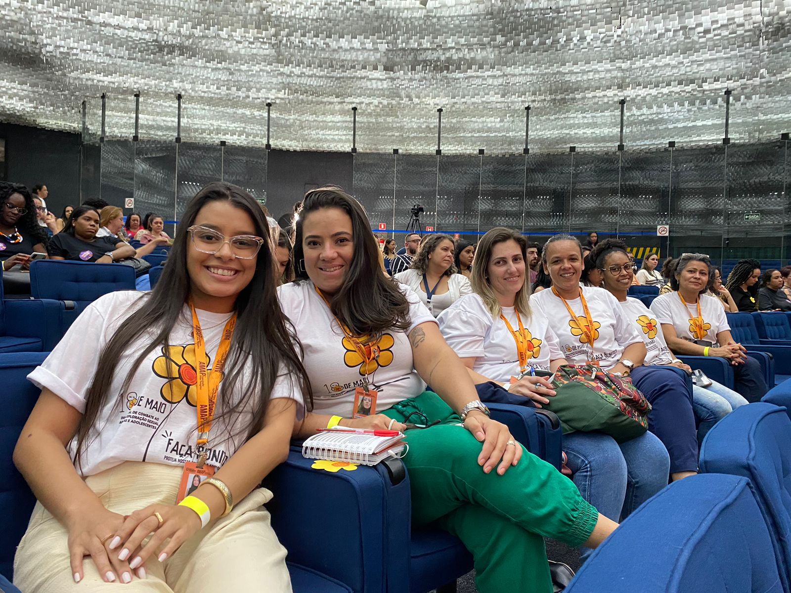 Na imagem aparecem seis mulheres sentadas em poltronas azuis, com uma camiseta branca com uma estampa de margarida laranja, da campanha 