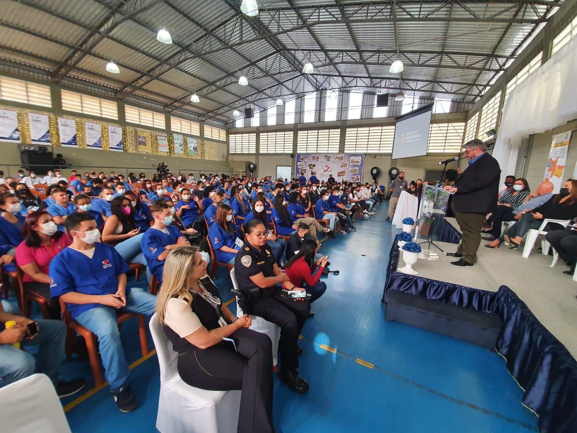 Na imagem aparece ao lado direito, o secretário adjunto da SMADS Décio Matos, e no lado esquerdo, pessoas sentadas viradas para um palco