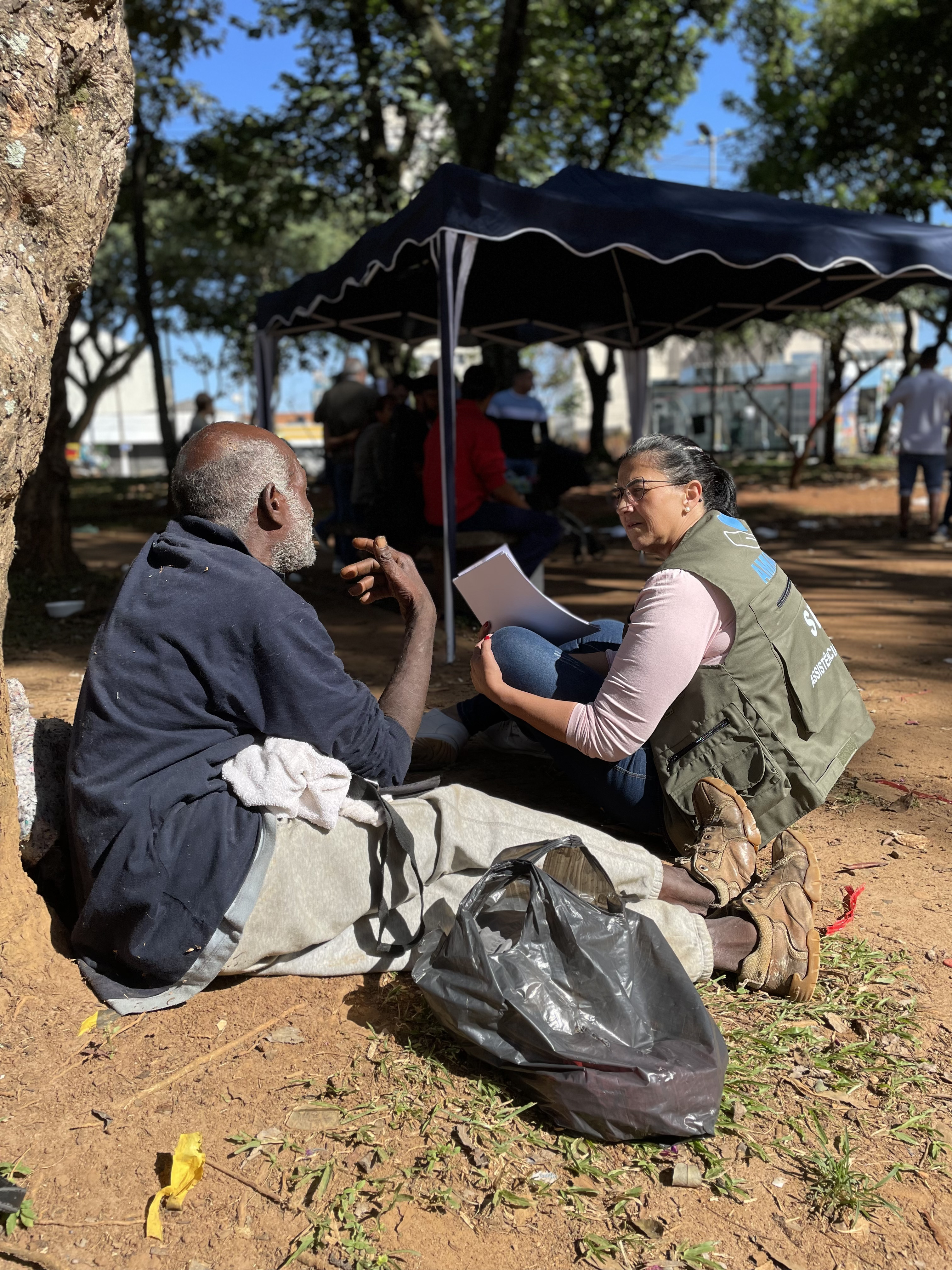 Na foto, um homem de calça cinza e camisa azul está sentado ao pé de uma árvore. Ele está conversando com uma mulher que veste rosa e jeans azulado, com o colete verde do Ampara SP. No fumdo, algumas pessoas estão embaixo de uma tenda azul.