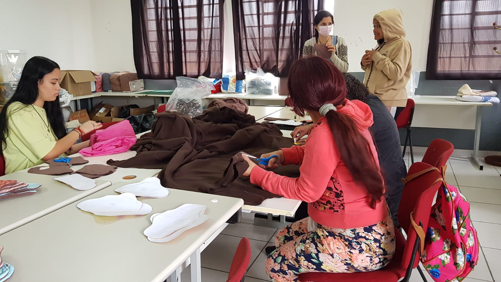 Três mulheres se sentam à mesa, cortando em tecido marrom os moldes dos absorventes. Em cima da mesa há os moldes de papel, tecido marrom, tecido rosa e tesouras. Ao fundo, há duas mulheres em pé conversando, enquanto uma delas segura um absorvente em suas mãos. 