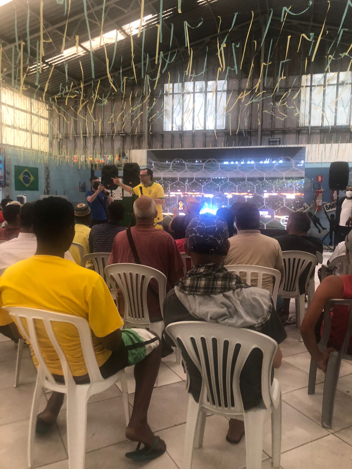 Algumas pessoas se sentam em cadeiras brancas, e à frente, um homem vestibndo uma camisa amarela da seleção fala no microfone. Um homem vestindo camiseta azul fotografa com o celular em mãos. É possível ver, ao fundo, um telão e três caixas autofalantes. No teto, se encontra penduradas fitas verde e amarelas e bandeiras de diversos países.
