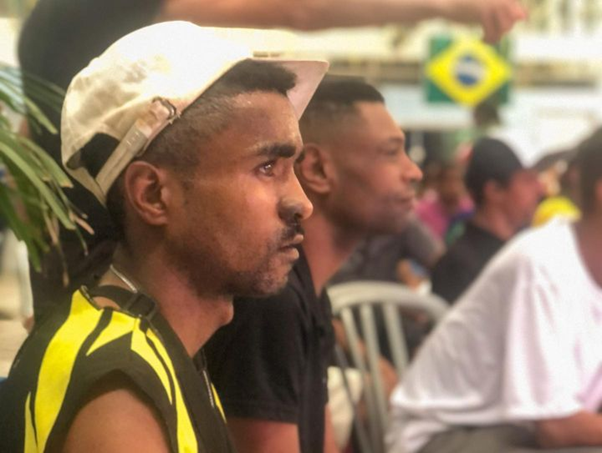 Homem de camisa regata e boné sentado em frente ao telão assistindo o jogo. 