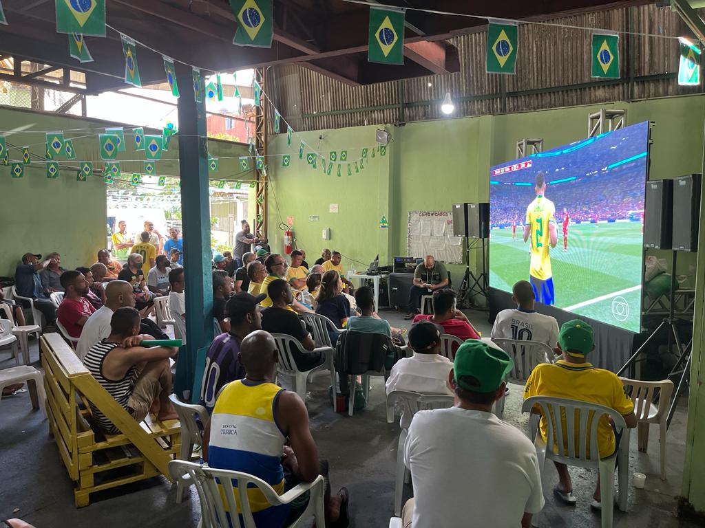 Na imagem aparece ao fundo um telão e bandeiras do Brasil, com pessoas, de costas para a imagem, sentadas assistindo o jogo. 
