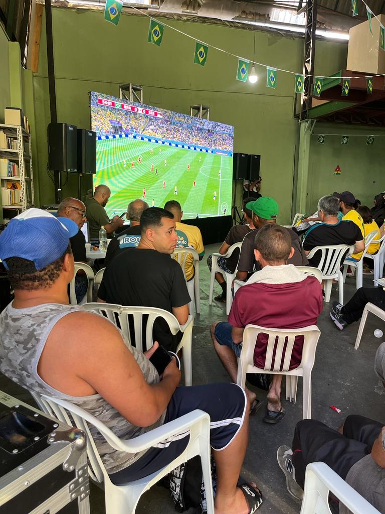 Homem de camisa regata e boné sentado em frente ao telão assistindo o jogo. 