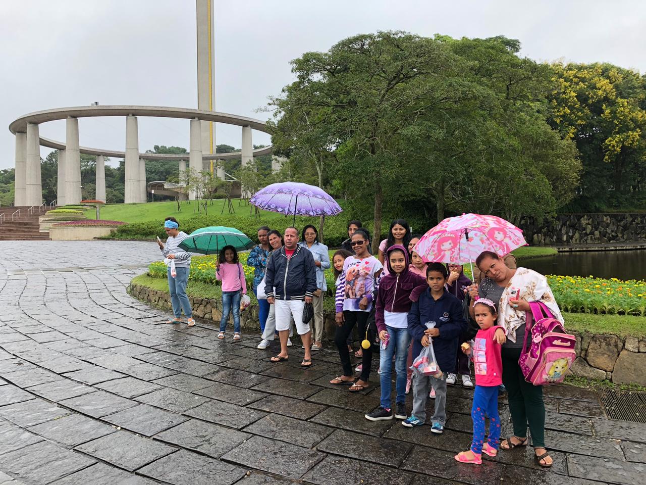 Homens, mulheres e seus filhos, em torno de 18 pessoas, posam para foto, algumas segurando guarda-chuvas, em jardim do Solo Sagrado de Guarapiranga