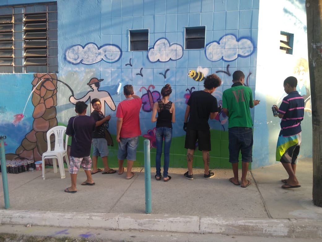 Sete jovens, uma menina e seis meninos grafitam parede externa do serviço. Um deles está olhando em direção à câmera. Um cenário com abelhas, pássaros, nuvens e grama verde está sendo pintado