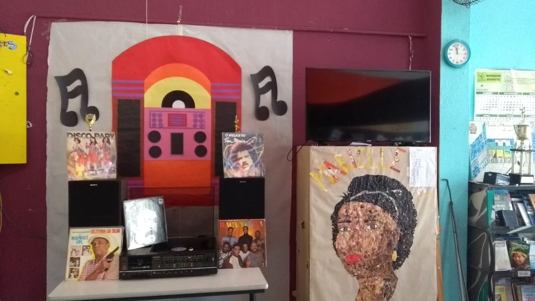 Exposição de vitrola e capas de discos de vinil com desenho colorido de vitrola e cifras musicais pregados na parede em ambiente enfeitado para a festa retrô do serviço