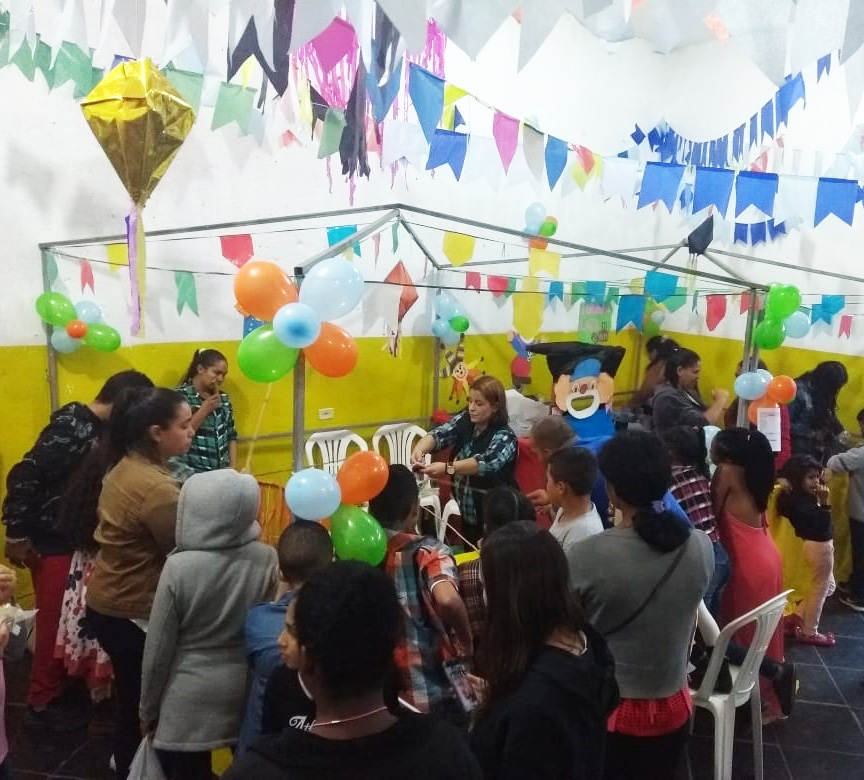 Crianças e adultos, em pé, ao redor de mesa branca de plástico, em salão do serviço, com bandeirolas coloridas no teto