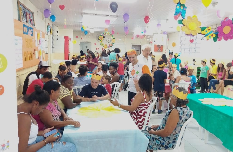 Conviventes de várias idades sentados em mesas de plástico brancas em sala com balões e flores coloridas de papel pendendo do teto. Alguns conviventes estão com coroa amarela de papel na cabeça