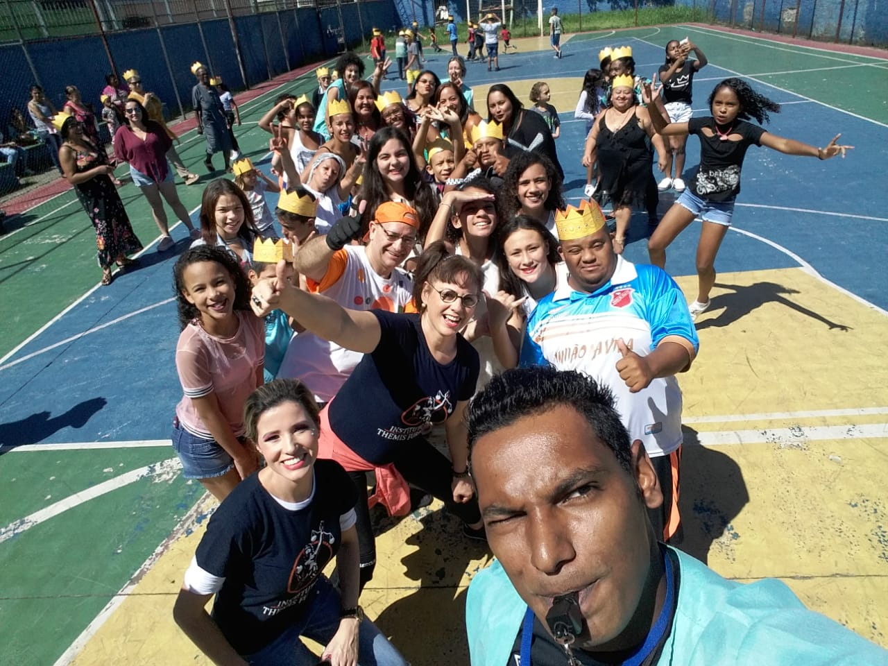 Conviventes fazem foto em selfie em quadra esportiva com instrutor à frente com camisa verde clara e apito na boca