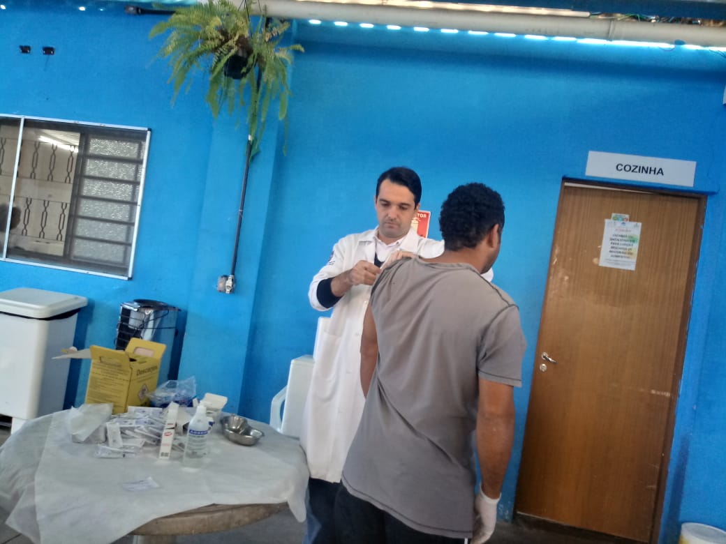 Convivente está em pé sendo vacinado por profissional da saúde, que usa jaleco branco, em parte externa do serviço
