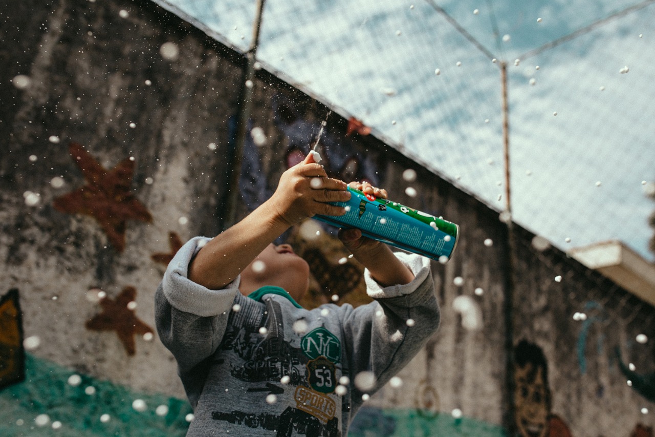 Menino brinca com espuma enquanto olha para cima sorrindo em pátio de esportes com parede grafitada e tela de proteção