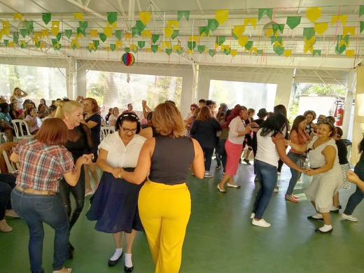 Mulheres dançam em pares em salão com teto branco de metal e bandeirolas verdes e amarelas no alto