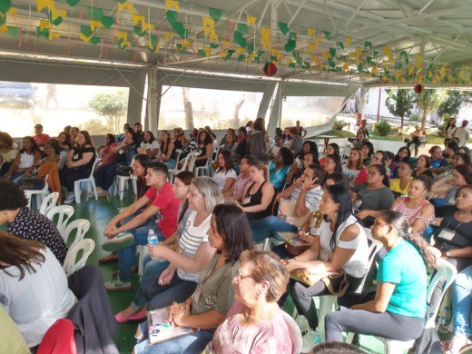 Plateia de mulheres e homens sentados em cadeiras de plástico brancas em local com teto branco de metal e bandeirolas verdes e amarelas no alto