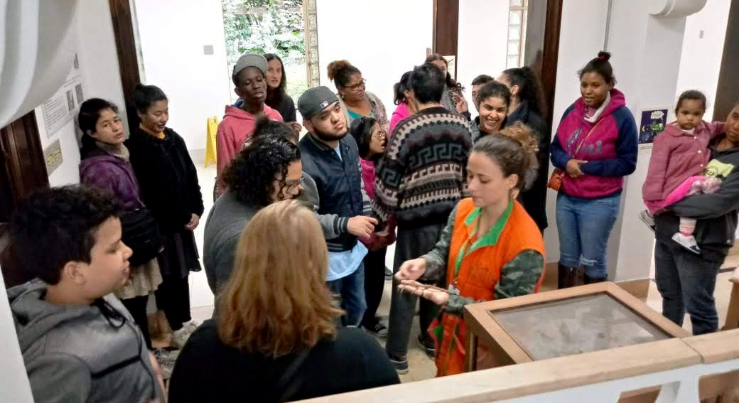 Cerca de dezenove conviventes em sala de exposição, prestando atenção na fala de monitora vestida com colete laranja