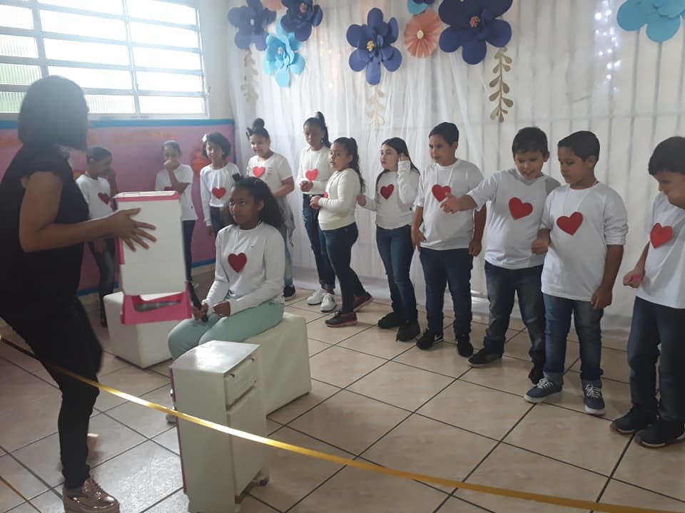 Doze crianças com camisa branca e coração vermelho no meio. Dez estão em pé e duas sentadas enquanto orientadora está a frente delas também em pé