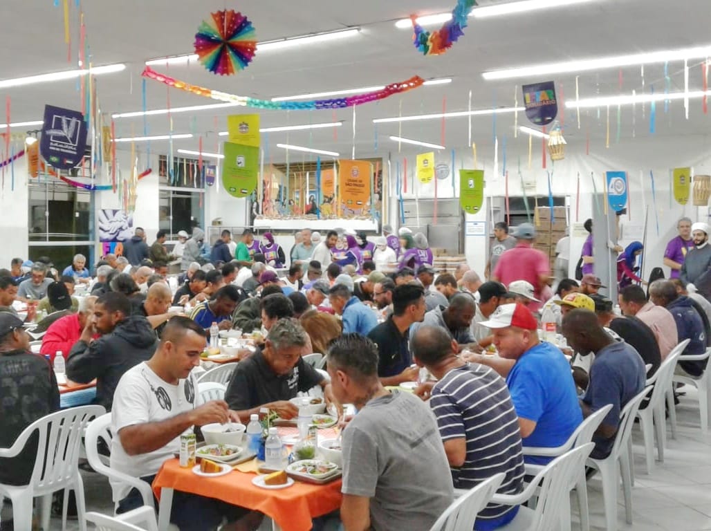 Conviventes jantam em mesas brancas de plástico em refeitório enfeitado com bandeirolas e fitas coloridas