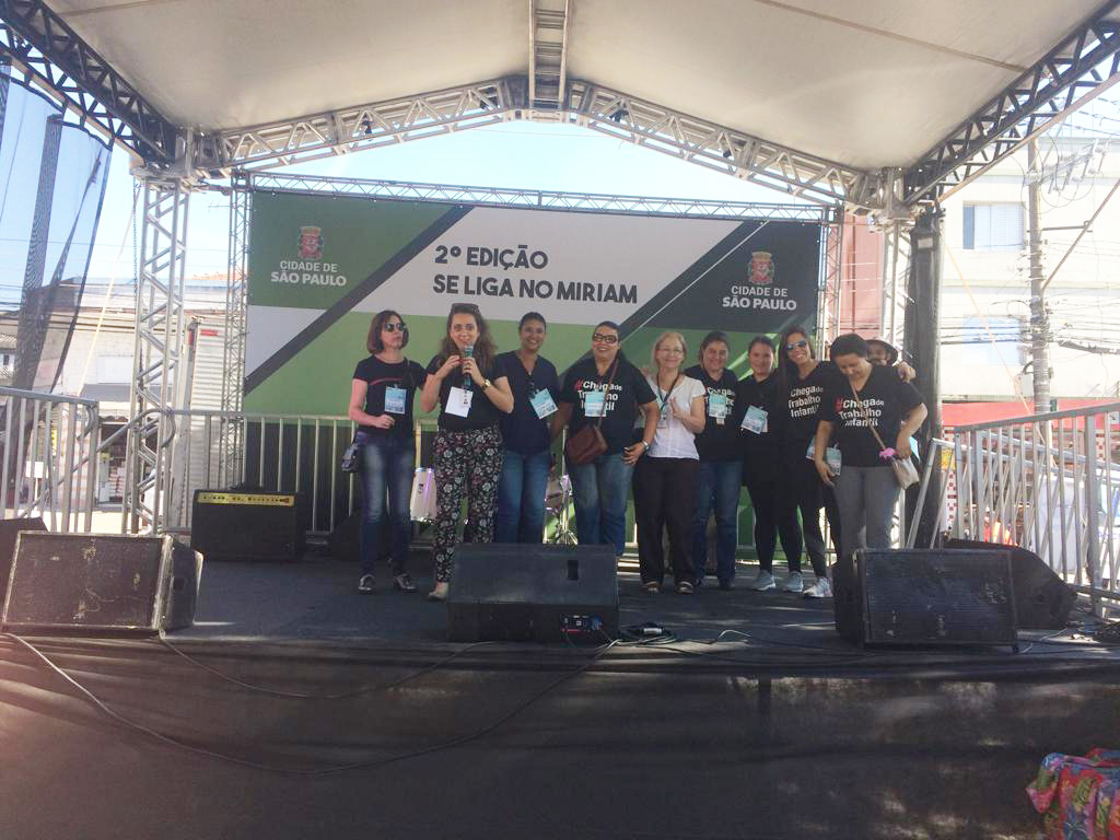Nove mulheres em pé, no palco do evento. No fundo há um painel verde e branco no qual se lê segunda edição do se liga no Miriam e o brasão da Prefeitura da Cidade de São Paulo no canto esquerdo e também no canto direito