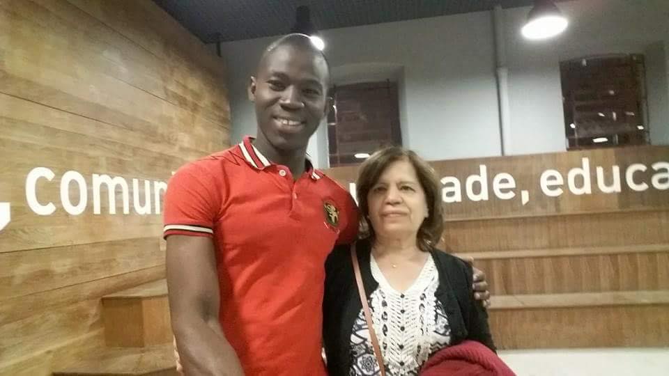 Abdul Guibila e a professora Sonia Altomar, abraçados. Ele usa uma camisa polo vermelha e sorri. Ela está com camisa branca e uma blusa de frio preta, com expressão séria. No fundo há uma arquibancada de madeira em toda a extensão do ambiente