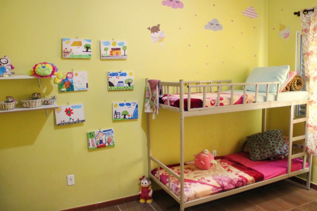 Um quarto com beliche, travesseiros, roupas de cama, brinquedos e paredes amarelas decoradas com nuvens rosas e quadros de temática infantil pendurados nas paredes