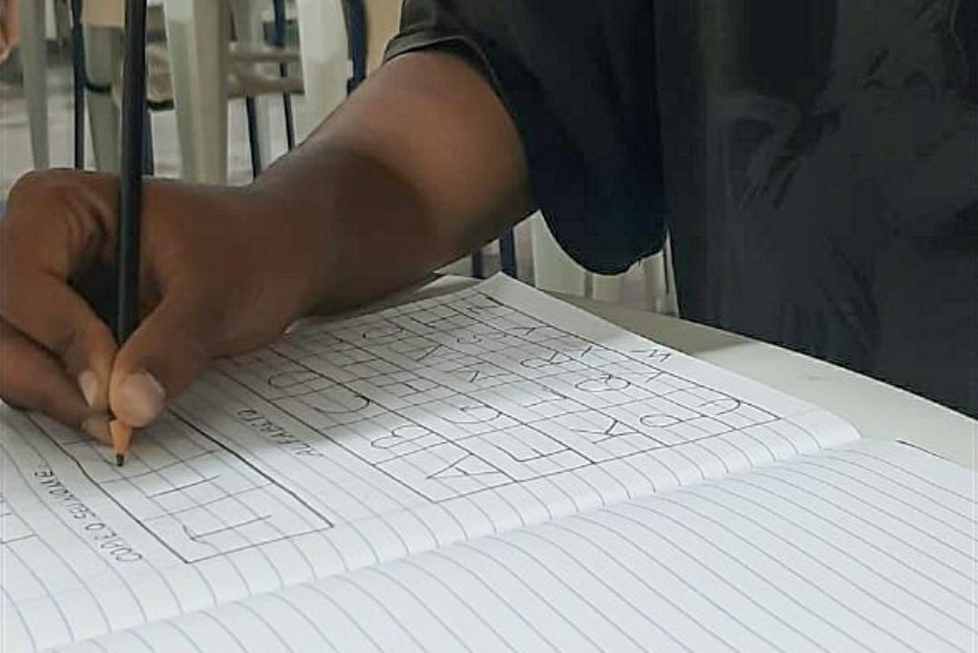 No centro da foto está o caderno do acolhido com as atividades e na parte superior está a mão dele com um lápis realizando os exercícios de aula