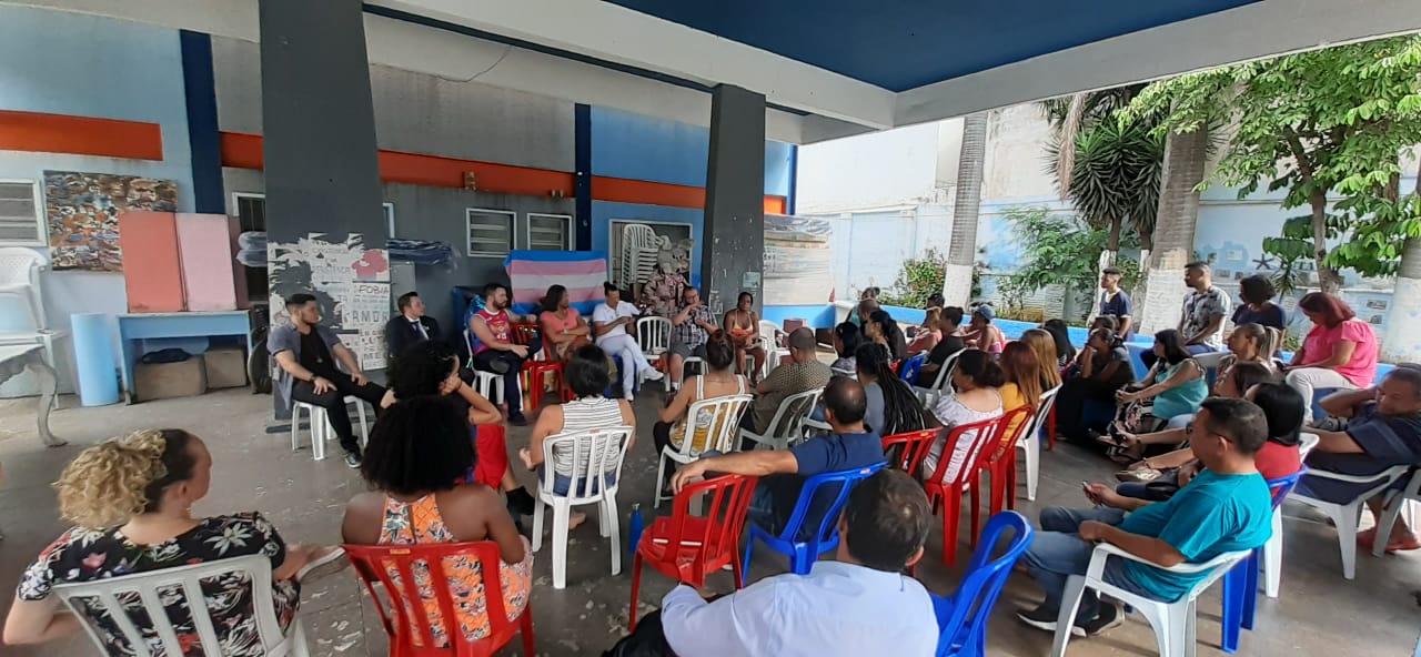 :  Dezenas de pessoas estão num espaço externo, sentadas em cadeiras e conversando.