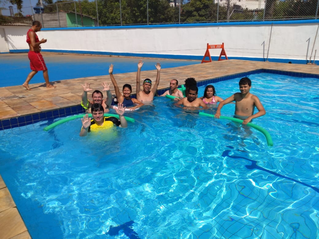 Nove pessoas estão dentro de uma piscina. Todas elas olham para a foto. Alguns seguram objetos de natação.