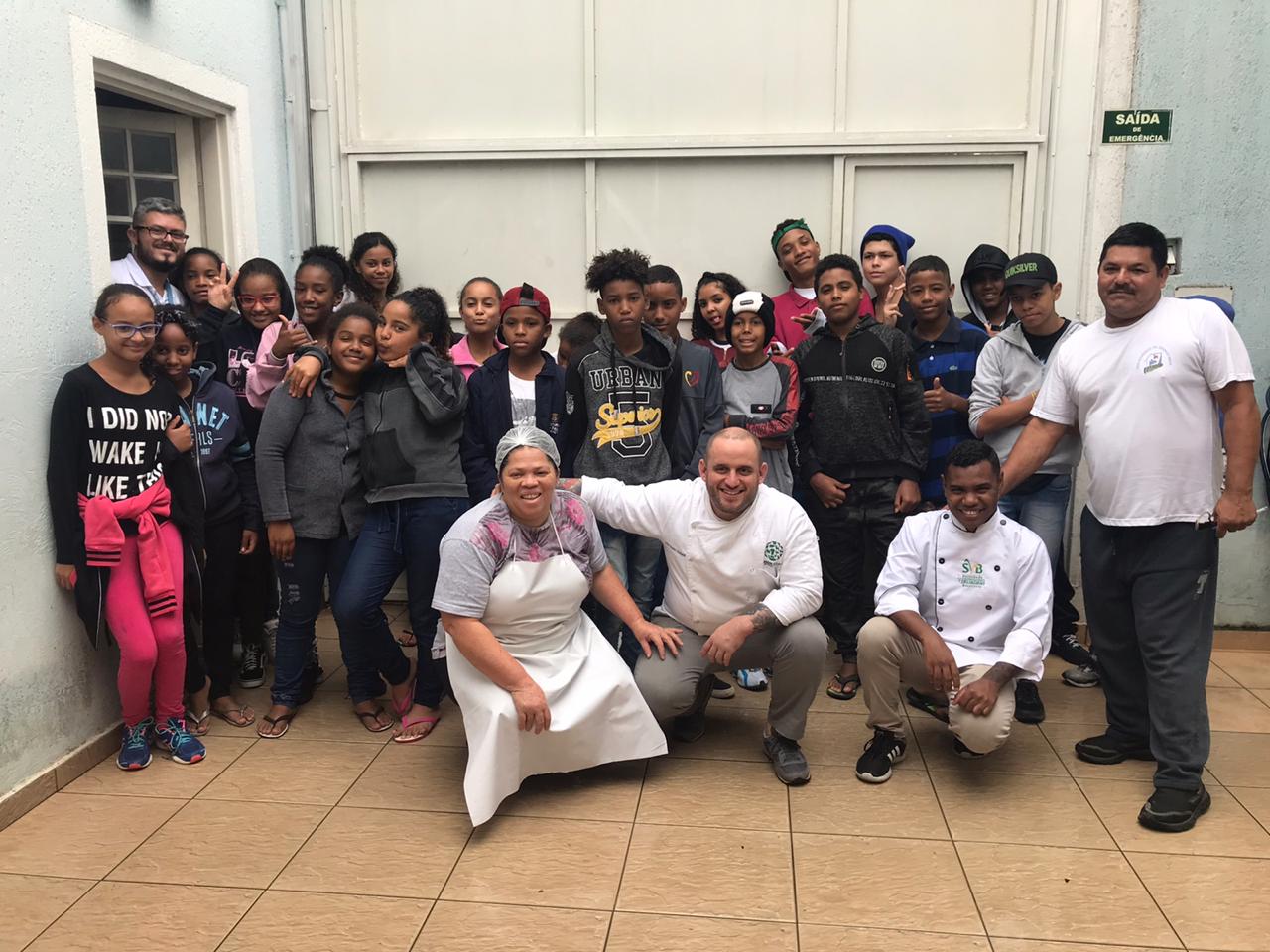 Dezenas de crianças estão reuniadas posando para a foto. Na frente deles, há uma mulher e três homens usando roupas de cozinheiro.