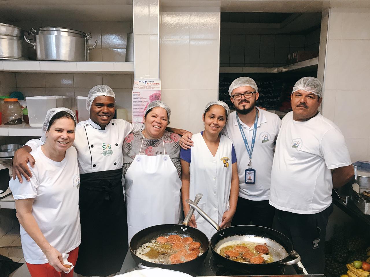 Três homens e três mulheres estão abraçados, de pé, dentro de uma cozinha. Todos olham e fazem pose para a foto.