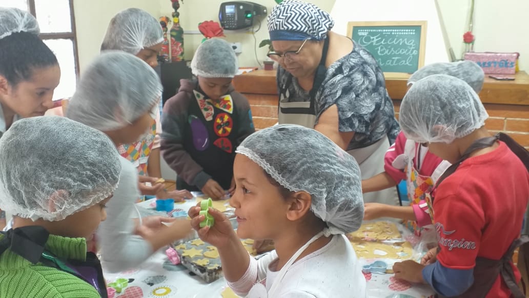Nove crianças, acompanhadas de uma senhora , estão preparando biscoitos dentro de uma cozinha. Todos usam touca e avental.