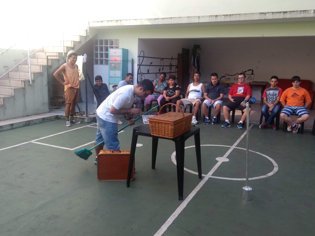 Dentro de uma quadra de futebol de salão nove pessoas estão sentadas em cadeiras. No centro da quadra, há um homem com uma vassoura entre as pernas ao lado de uma mesa de plástico preta com uma cesta marrom em cima. Ao lado da mesa, no chão, há uma maleta bege.